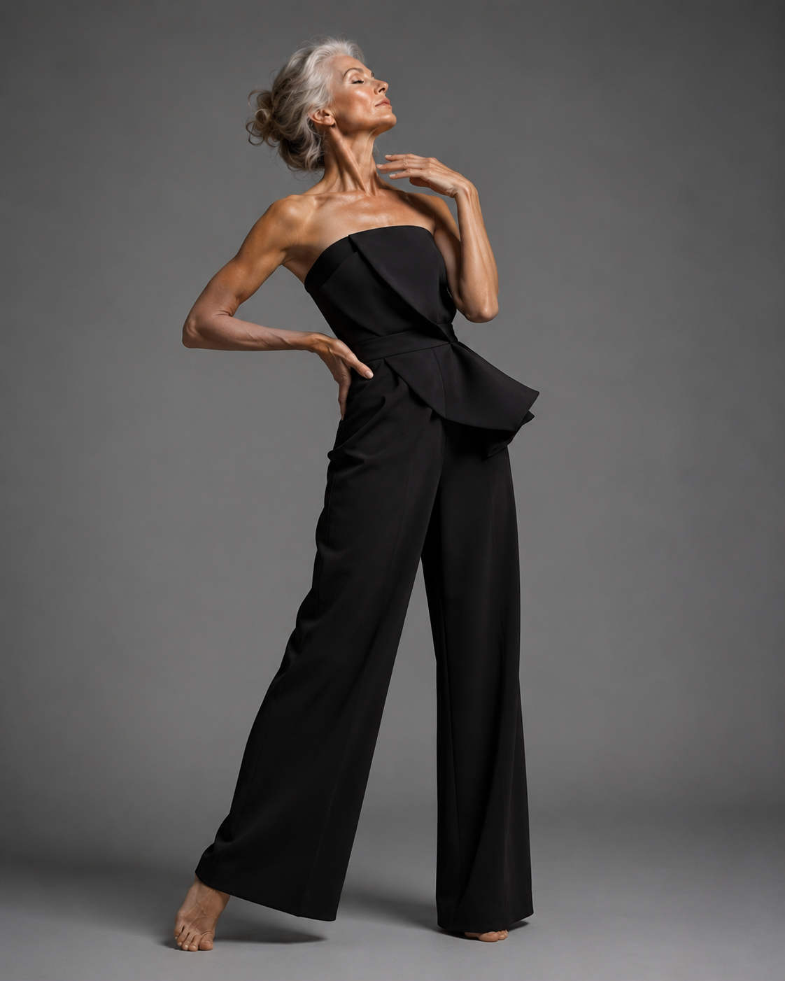 mature silver-haired woman, grey studio backdrop in black strapless peplum jumpsuit, poised profile, hand at neck, chin lifted