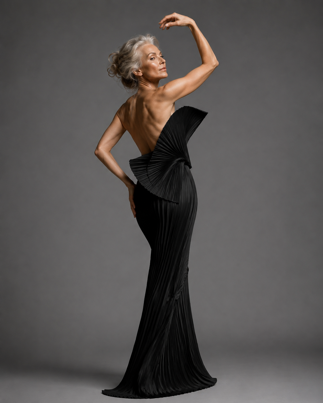 mature silver-haired woman, grey studio backdrop in sculptural black gown with bow back, back view, arm curved overhead