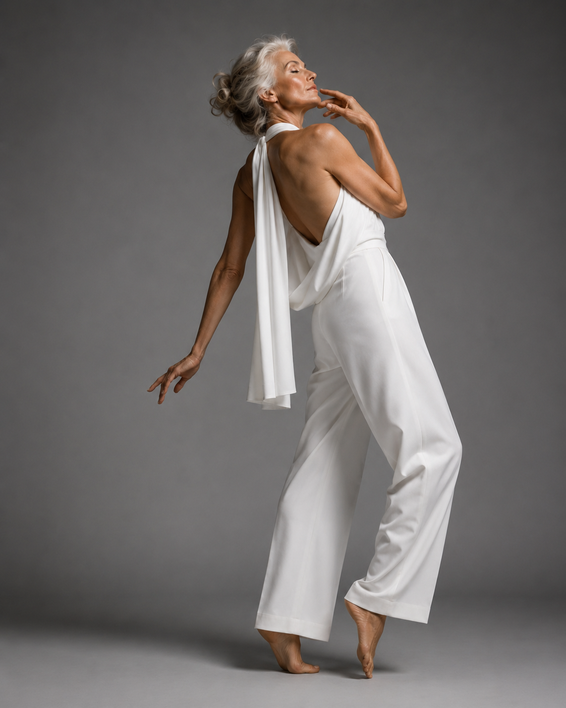 mature silver-haired woman, grey studio backdrop in white draped halter jumpsuit, back view, arm raised