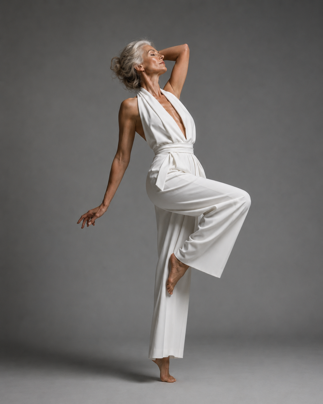 mature silver-haired woman, grey studio backdrop in white halter jumpsuit, leg raised behind, arm overhead